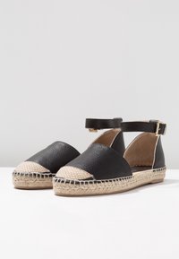 Espadrilles de cuero negro con suela de yute natural, punta de almendra y tirante al tobillo con detalle de hebilla dorada.
