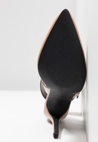 Chaussure à talon stiletto beige à bout pointu avec semelle noire texturée, positionnée verticalement contre une surface blanche.