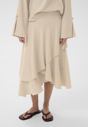 Beige layered midi skirt paired with a matching long-sleeve top featuring wide cuffs with gold buttons, worn with dark brown flat sandals.