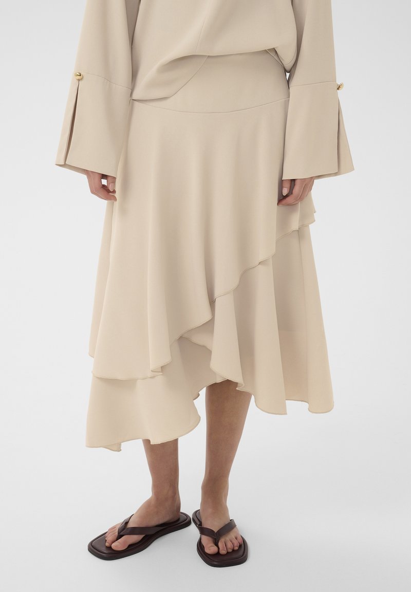 Jupe midi beige à plusieurs couches associée à un haut à manches longues assorti, doté de larges poignets avec des boutons dorés, portée avec des sandales plates marron foncé.