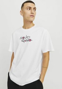 Hvid bomulds t-shirt med "SQUID GAME" trykt i sort og pink. Korte ærmer, rund halsudskæring, normal pasform med en glat overflade.