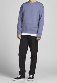 Felpa azzurra chiara con collo rotondo, polsini e orlo a coste, indossata sopra una camicia bianca; abbinata a pantaloni neri e sneakers nere con lacci bianchi.