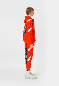 Joven con sudadera con capucha y pantalones naranjas brillantes con diseños de alas y etiquetas negras, de perfil y con botas blancas con cordones.