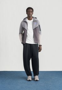 Pelēka un bēša krāsas kapuce ar rāvējslēdzēju un kapuci, ar zilu logotipu. Kombinēts ar melnām, vaļīgām biksēm un gaiši pelēkām sporta apaviem.