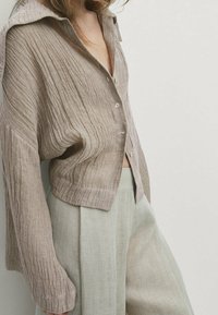 Een button-up overhemd in lichtbruin textuurstof met verticale ribbels, gecombineerd met lichtbeige wijde broekspijpen, die een ontspannen pasvorm accentueert.