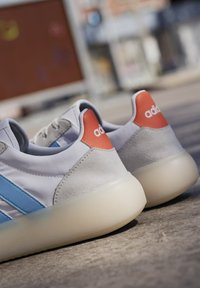 Vita sneakers med blåa ränder och oranga häldetaljer. Tillverkade av släthet tyg och mocka, med en transparent gummisula.