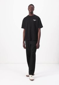 Black oversized t-shirt with white logo, paired with black trousers and light-colored sneakers. Simple, relaxed fit design.