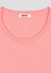 Camiseta de algodón rosa con cuello redondo y una etiqueta que dice "JOTT" en rojo. La tela parece suave con una textura sutil.