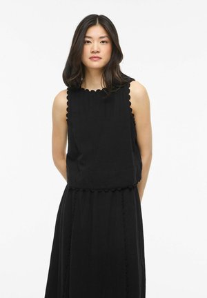 Jeune femme aux cheveux mi-longs, vêtue d'une robe noire sans manches aux bords festonnés, debout devant un fond blanc uni.