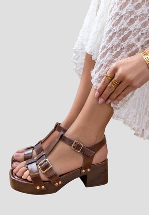 Pieds portant des sandales à talons en cuir marron avec des boucles dorées, reposant sur une jupe en dentelle blanche, main avec des bagues et un bracelet en or touchant la cheville.