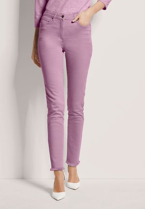 Femme portant un jean slim rose avec des ourlets effilochés et des talons blancs à bout pointu, main dans la poche, debout contre un mur clair.