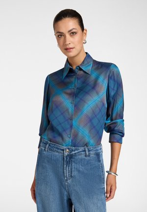 Donna con capelli scuri con una camicia a quadri blu e viola abbottonata e jeans blu, in piedi con una mano rilassata lungo il fianco.