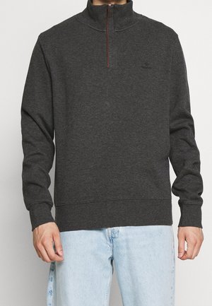 Tummanharmaa vetoketjupullovari, jossa on korkea kaulus, puoliväli-vetoketju ja joustavat hihansuut. Pehmeä rakenne ja hillitty logo rinnassa.