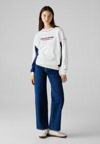 Felpa bianca con accenti rossi e blu marino e testo "RED BULL RACING". Abbinata a jeans a gamba larga blu e sneakers bianche con suola marrone.