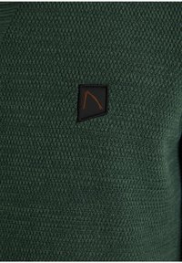 Dunkelgrüner Strickpullover mit strukturierter Oberfläche und einem kleinen schwarzen Etikett auf der Brust, das eine orangefarbene Umrandung eines Dreiecks zeigt.