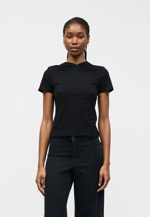 Γυναίκα που στέκεται και κοιτάζει προς τα εμπρός, φορώντας ένα απλό μαύρο κοντομάνικο μπλουζάκι και μαύρο παντελόνι, μπροστά σε ένα ανοιχτό γκρι φόντο.