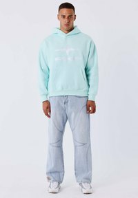 Haut à capuche bleu clair avec poche avant et logo blanc, associé à un jean en denim clair avec des coutures courbées et un patch logo. Baskets blanches.