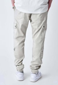 Persona che indossa pantaloni cargo beige chiaro con polsini elasticizzati, diverse tasche con bottoni, sneakers bianche e una camicia bianca, con le mani nelle tasche.