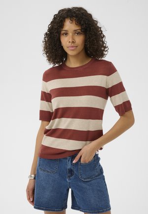 Kvinde med krøllet hår iført en kortærmet, rust- og beige-stribet top og blå cowboyshorts, stående med den ene hånd i lommen.