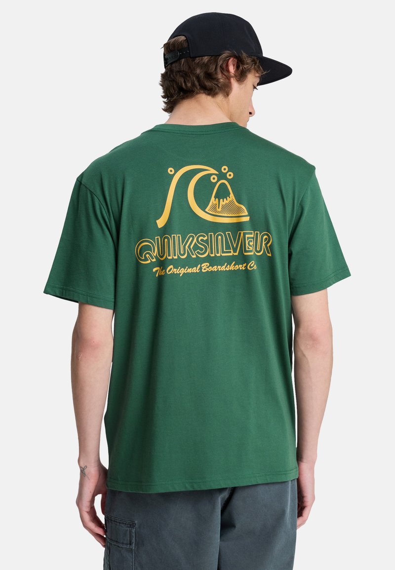 Jeune homme portant un t-shirt vert Quiksilver avec un logo jaune et du texte au dos, une casquette noire et un short cargo foncé, vu de dos.