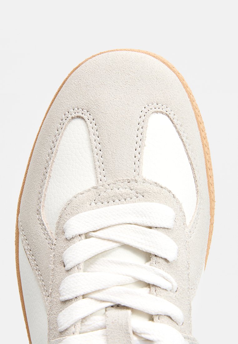 Primo piano di una punta di sneaker bianca e grigia chiara con lacci bianchi e suola in gomma beige, che mostra materiali in suede e pelle.