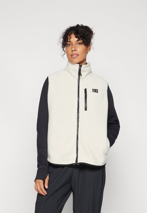 Femme aux cheveux bouclés portant une veste polaire blanche zippée par-dessus un t-shirt noir à manches longues et un pantalon noir, debout devant un fond uni.