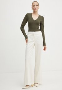 Top fitted verde oliva a maniche lunghe con scollatura a V; abbinato a pantaloni bianchi con gamba larga; scarpe bianche a punta con dettagli dorati.