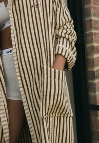 Persoon die een bruin-wit gestreepte badjas draagt met de tekst "self." geborduurd, hand in de zak, naast een andere persoon in witte "self." onderkleding.