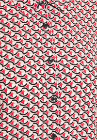 Chemise à motifs avec un fond blanc ornée de motifs noirs, rouges et beiges. Des boutons noirs sont disposés verticalement sur le devant.