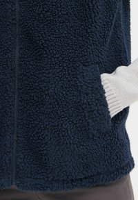 Marinblå fleecejacka med en texturerad yta, med dragkedja och sidofickor. Ljusgrå ribbade muddarna är synliga.