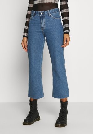 Person trägt hoch taillierte, blaue, kurz geschnittene Jeans mit rohen Säumen, kombiniert mit schwarzen Schnür-Combat-Stiefeln und einem schwarz-weißen Langarmshirt.