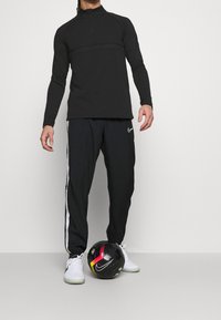 Schwarzes Langarm-Sportoberteil mit Reißverschluss, kombiniert mit schwarzen Hosen mit weißen Seitenstreifen, und einem schwarzen Fußball auf dem Boden.
