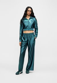 Σετ φόρμας από σατέν σε τιρκουάζ με κοντό μπουφάν με φερμουάρ και παντελόνια με ελαστική μέση. Διαθέτει λογότυπο PUMA σε ανοιχτό τιρκουάζ. Το μοντέλο φοράει ταιριαστά τιρκουάζ αθλητικά παπούτσια.