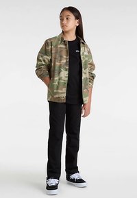 Veste camouflage vert et beige, t-shirt noir, pantalon noir, et baskets noires à semelles blanches. Le modèle se tient les mains dans les poches.