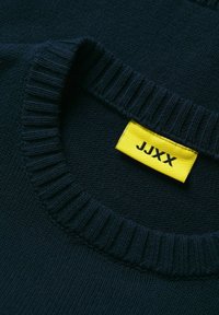 Jūras spēku džemperis ar ribu apkakli. Iezīmēts ar dzeltenu etiķeti ar nosaukumu "JJXX." Tuvplāns, kas uzsver tekstūru un šuvju detaļas.