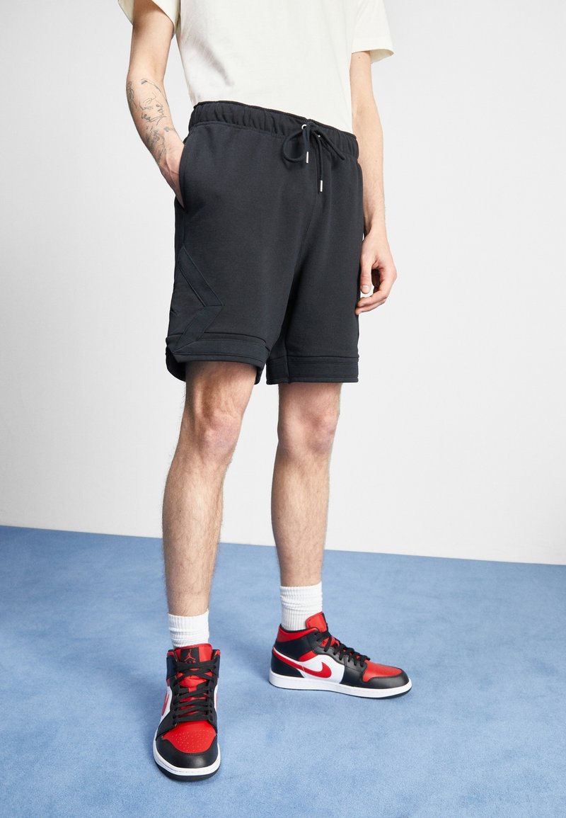 Svarta sportshorts med elastisk midja och draremsor. Har snedställda sidofickor och distinkta fålldetaljer. Bärs med röda och svarta sneakers.