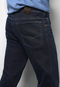 Dark blue denim jeans with a straight cut, featuring a brown leather patch at the back waistband and two back pockets with contrasting stitching.