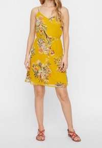 Robe fleurie jaune à fines bretelles, taille cintrée et ourlet fluide. Présente des imprimés floraux multicolores et est associée à des sandales rouges à fines lanières.