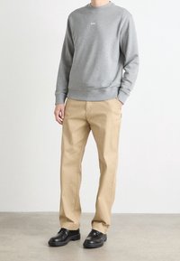 Hombre vistiendo una camisa de manga larga gris con el texto "BOSS", pantalones beige y zapatos negros, de pie sobre un suelo liso frente a un fondo blanco.