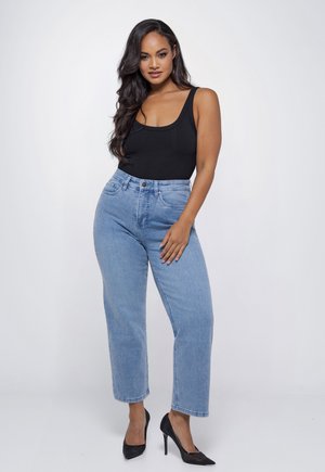 Vrouw staand, gekleed in een zwart mouwloos topje, lichtblauwe hoge taille jeans en zwarte hoge hakken, tegen een effen achtergrond.