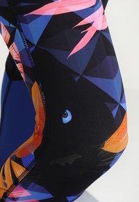 Kolorowe legginsy z geometrycznym wzorem w odcieniach czerni, niebieskiego i pomarańczowego; zawierają graficzne elementy ryb i liści.