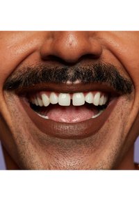 Närbild av en leende mun med vita tänder, bruna läppar och en framträdande mustasch. Hudens struktur ser slät ut med fin hårväxt.