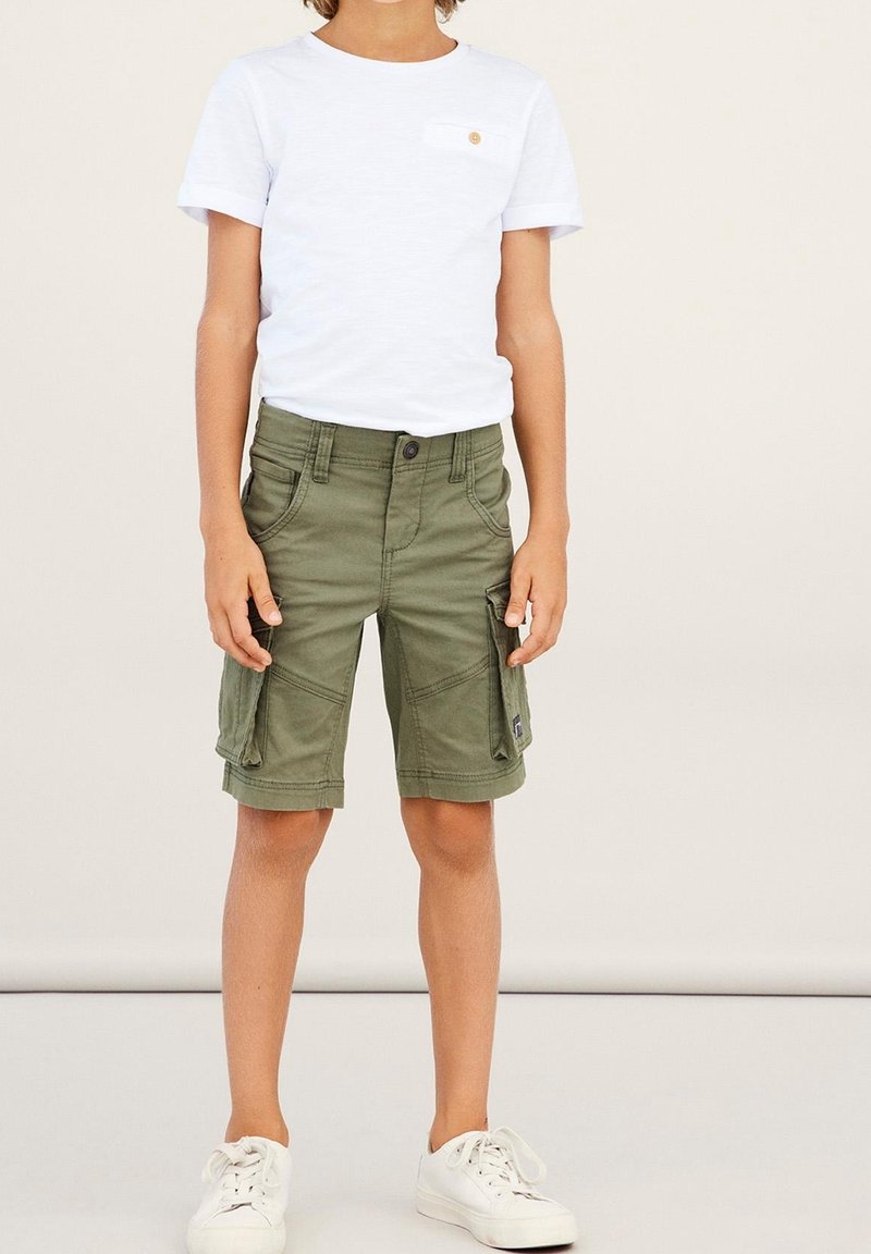 Enfant portant un t-shirt blanc à manches courtes, un short cargo vert olive avec poches latérales et des baskets blanches, debout devant un fond uni.
