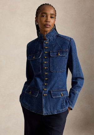 Kvinde med flettet hår iført en tætsiddende blå denimjakke med messingknapper, fire klaplommer, og står med hænder i lommerne.