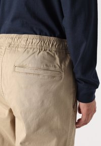 Beige cotton trousers with an elastic waistband, featuring a back pocket and smooth texture, displayed with a navy long-sleeve shirt.