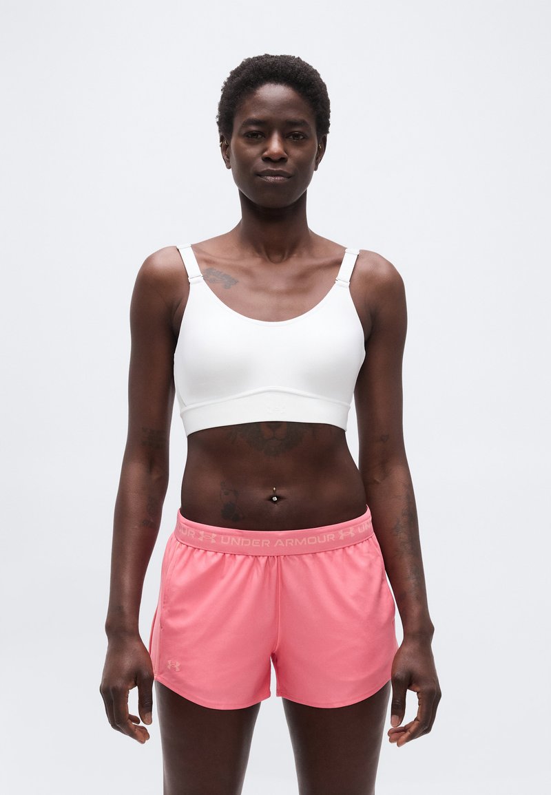 Αθλητική γυναίκα που φοράει άσπρο αθλητικό σουτιέν και ροζ σορτς Under Armour, στέκεται μπροστά σε λευκό φόντο.