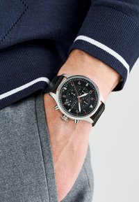 Black chronograph watch with silver stainless steel case, red accents, and black rubber strap. Features textured dial and multiple sub-dials.