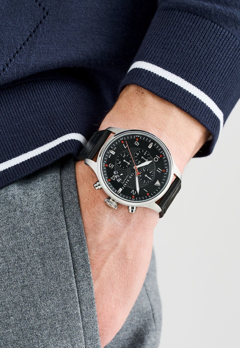 Black chronograph watch with silver stainless steel case, red accents, and black rubber strap. Features textured dial and multiple sub-dials.