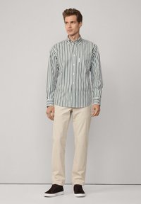 Camisa de botones a rayas en verde y blanco, de mangas largas, con cuello y bolsillo en el pecho; combinada con pantalones beige y calzado oscuro.