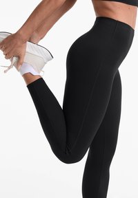 Colanți negri dintr-un material elastic, cu o talie înaltă și o textură netedă, asortați cu pantofi sport de culoare deschisă.